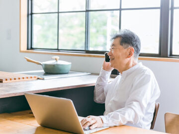 家のダイニングでスマートフォンで電話をする高齢男性